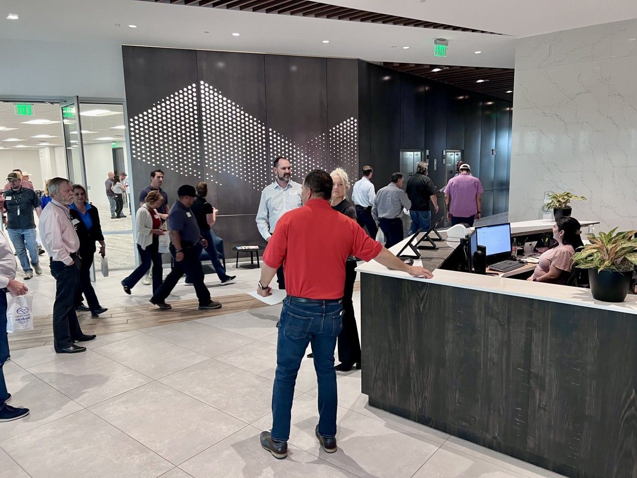 People in office lobby near a reception desk; some walking, some talking. Decorative wall in background.