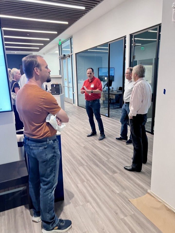 Group of people in an office hallway; a man speaks while others listen, one looks at a screen.