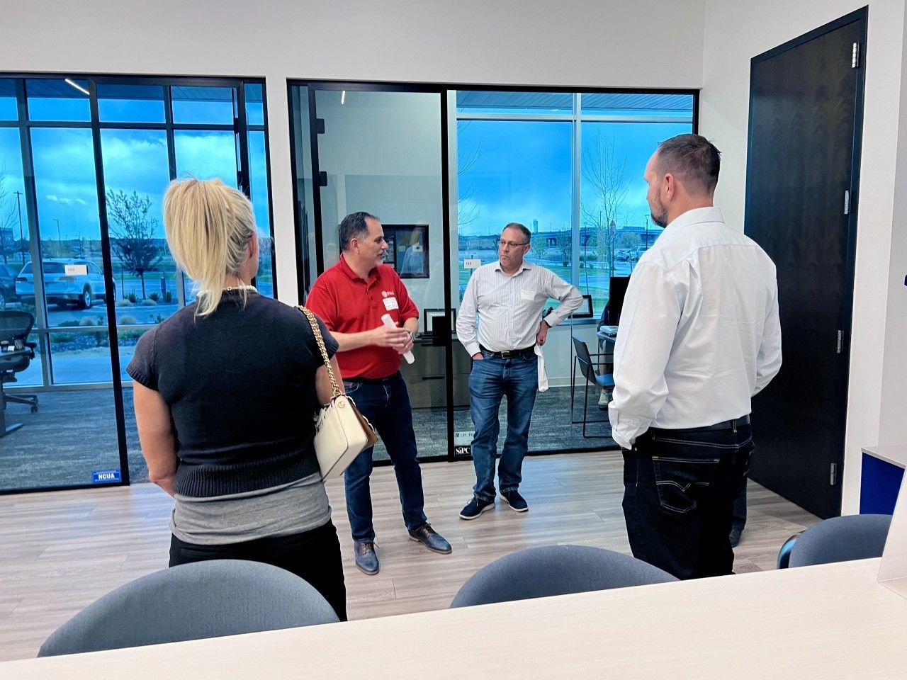 Four people in an office setting, conversing near large windows with a cloudy sky outside.