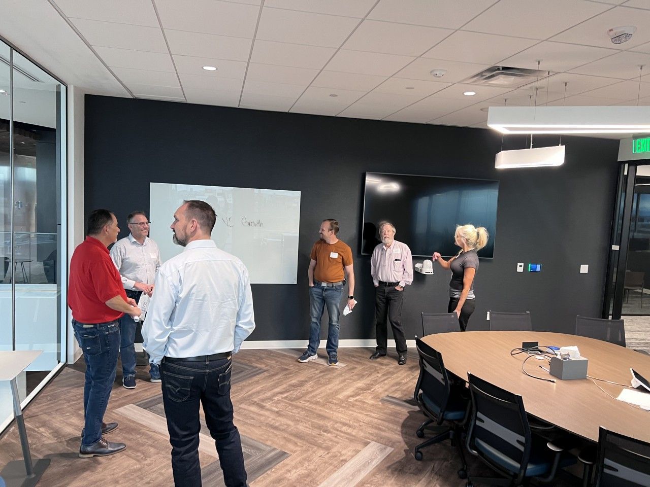 Group of people in a modern office, gathered around a whiteboard and conference table, discussing ideas.