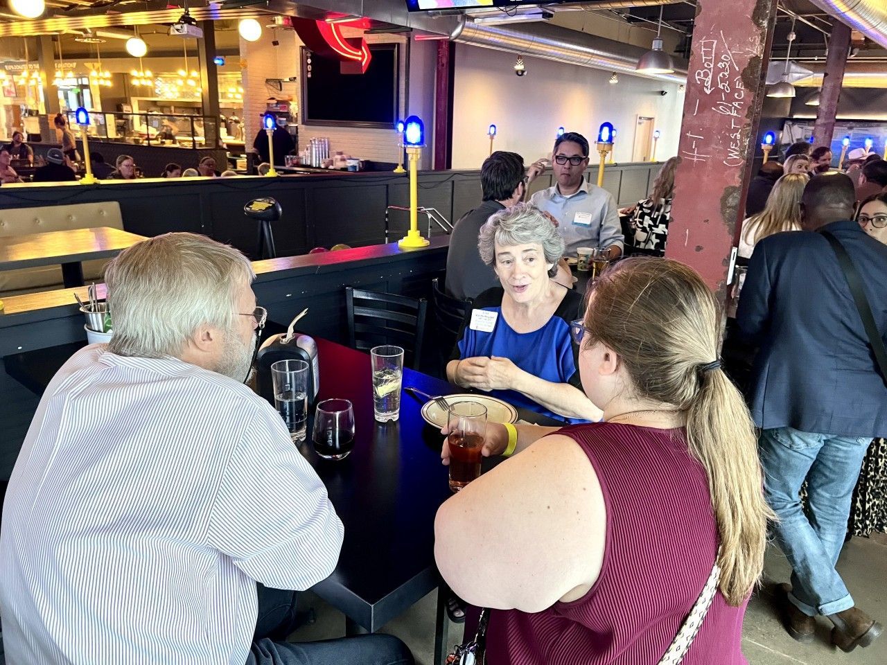 People at a table inside a bar, talking and drinking. Others are in the background.