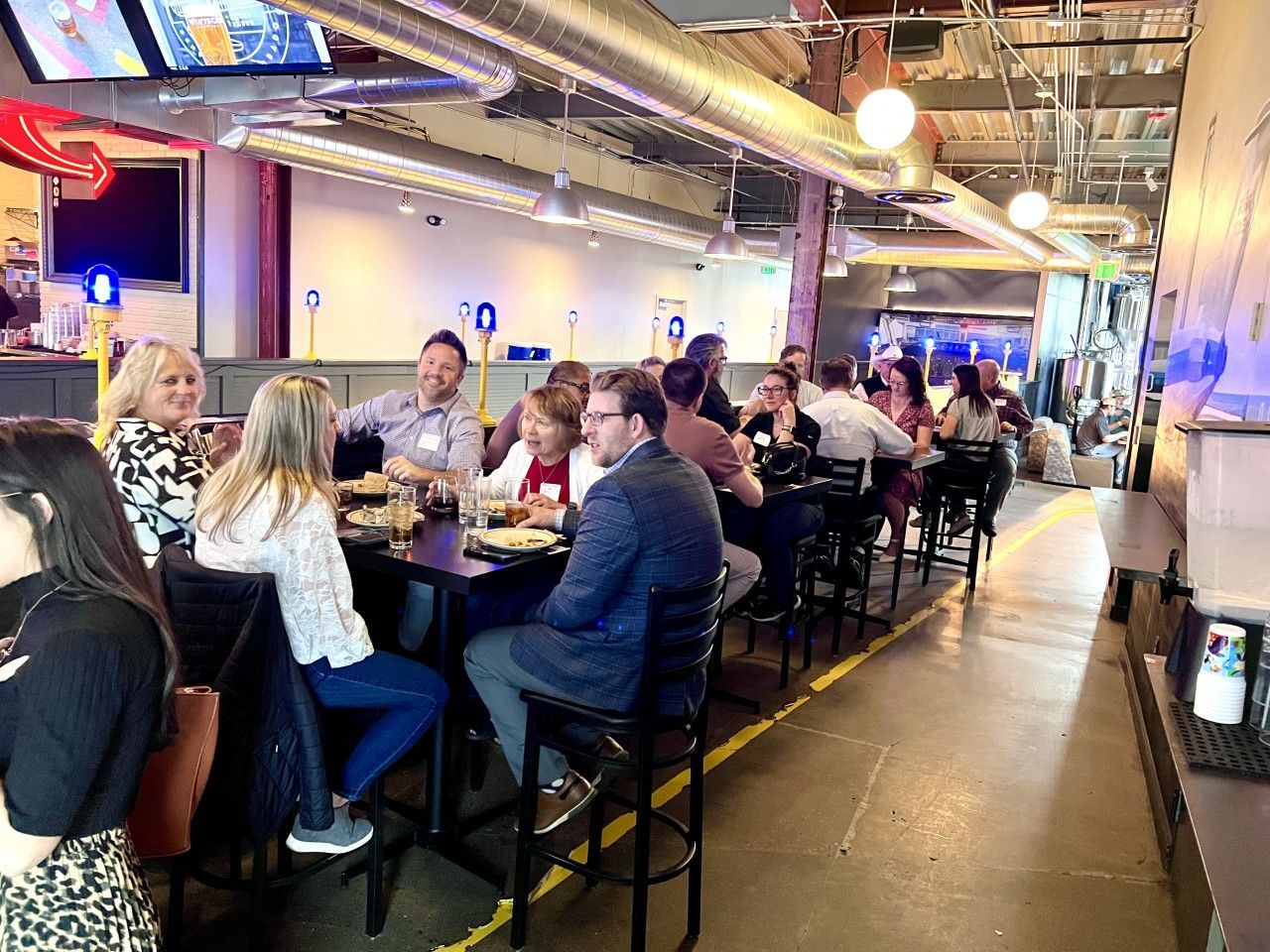 People seated at tables in a brightly lit restaurant, eating and talking. Long narrow space with high ceilings.