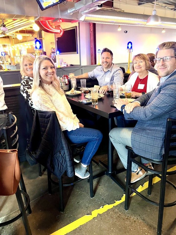 People at a table in a restaurant. Smiling woman in white top, others in business casual attire.