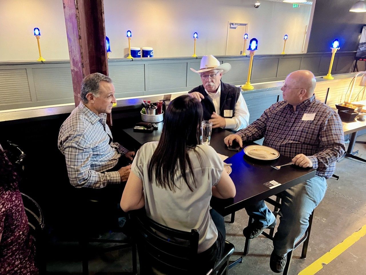 Four people seated at a table, conversing in a casual setting. One man wears a cowboy hat.