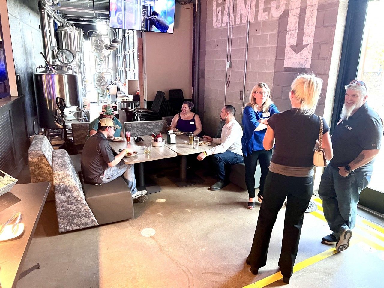 People socializing in a brewery, some seated at a table, others standing. Industrial setting with bar equipment visible.