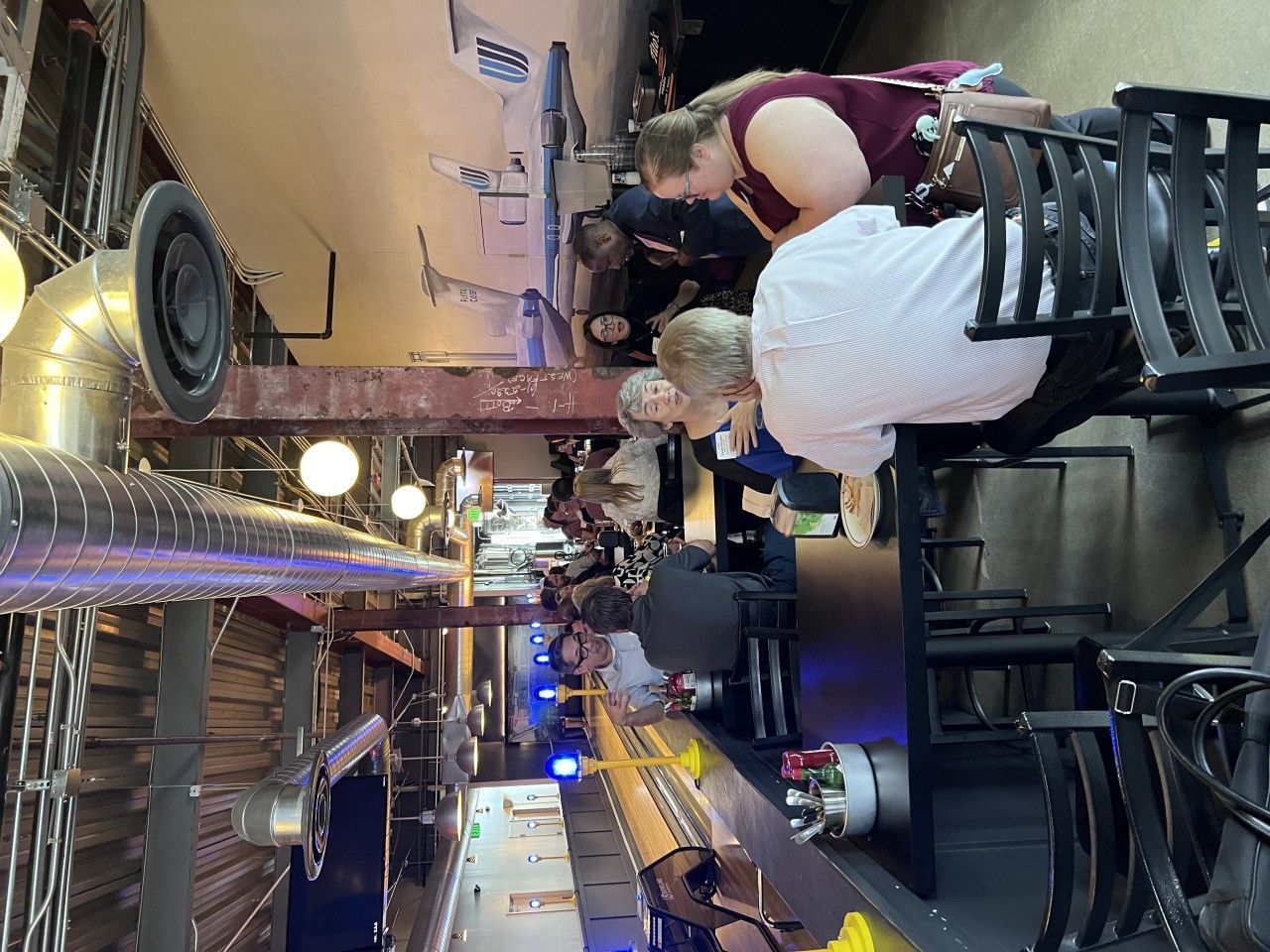 People at a bar, some seated, others standing. Long, narrow space with metalwork and overhead pipes.