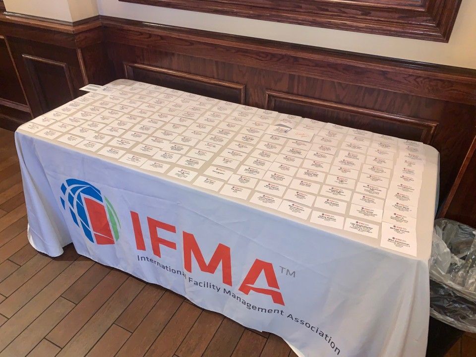 Table covered in a white IFMA tablecloth, with papers and a logo. Set in a room with wood paneling.