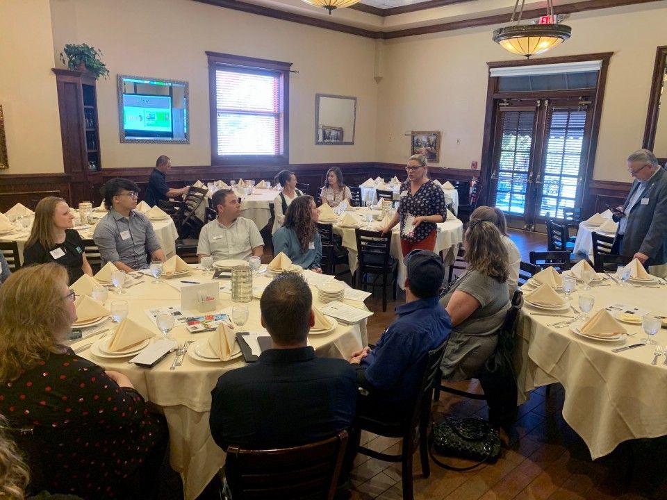 People seated at round tables in a restaurant, listening to a presentation.