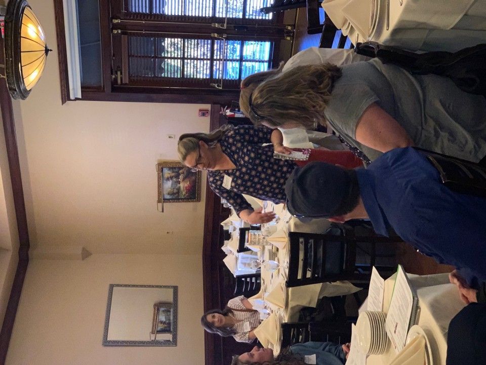 People gathered around a restaurant table. A woman in a patterned top leans in to serve. Formal setting.