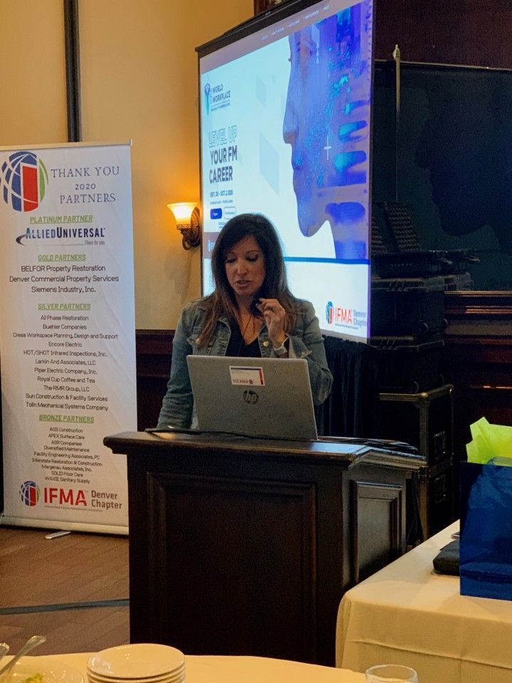 Woman presenting at a podium, a laptop open in front. A projected slide is behind her.