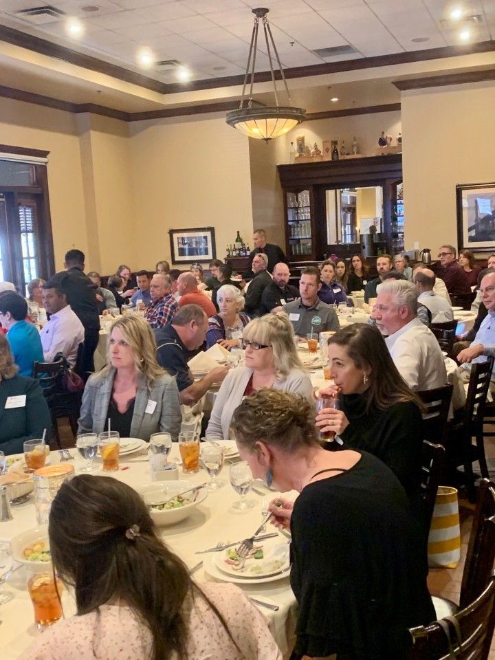 People at tables in a restaurant, attending an event. Tables are set with food and drinks.