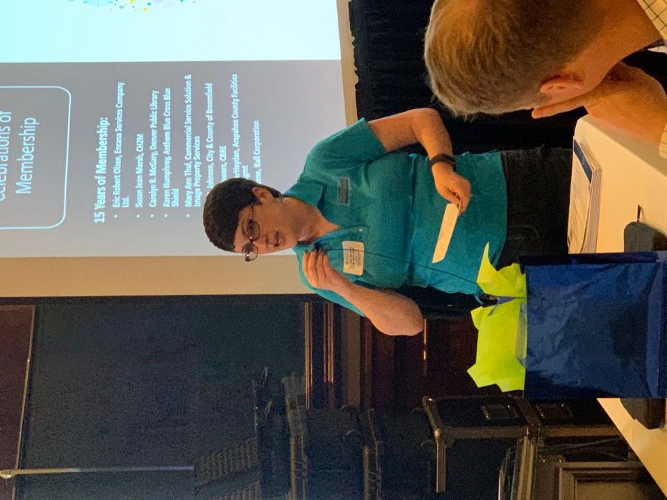 Woman in teal shirt speaks at a presentation, holding papers, with a screen and gift bag visible.