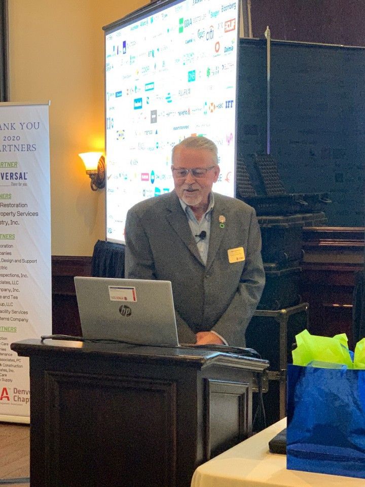 Man giving a presentation at a podium. A laptop sits on the podium. A screen displays logos behind him.