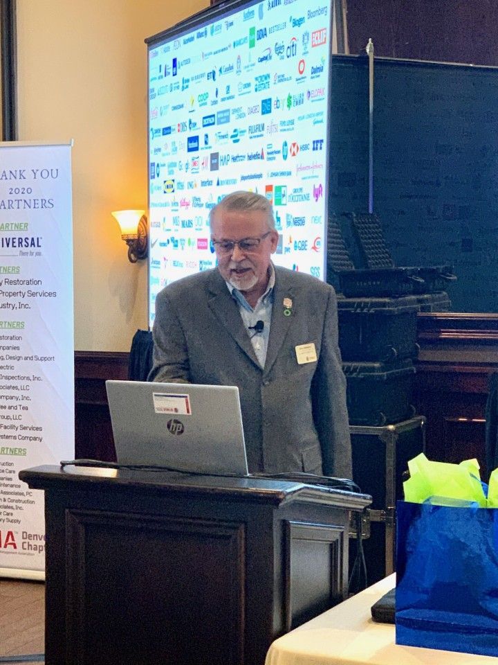 Man speaking at a podium, looking at a laptop. A screen behind him displays logos.