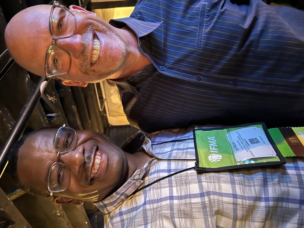 Two men smiling at camera, close together. One wearing a plaid shirt, the other blue stripes. Indoor setting.