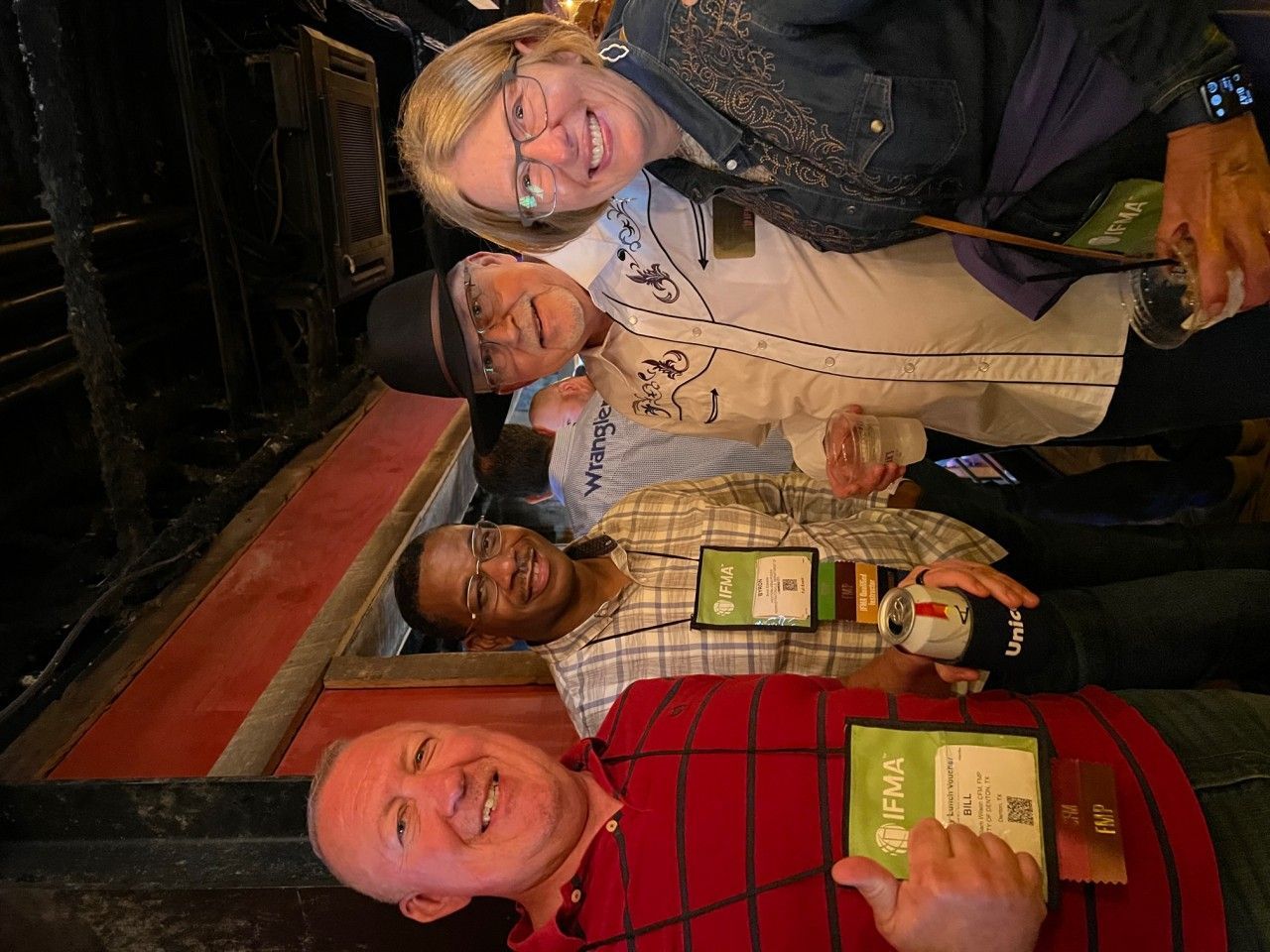 Four people smiling, holding drinks, near a stairwell. Three have tags. One person holds a wand.