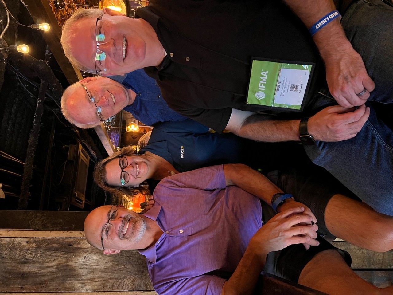 Four people smile, posing in a dimly lit indoor setting. One in purple shirt smiles, others wear glasses.