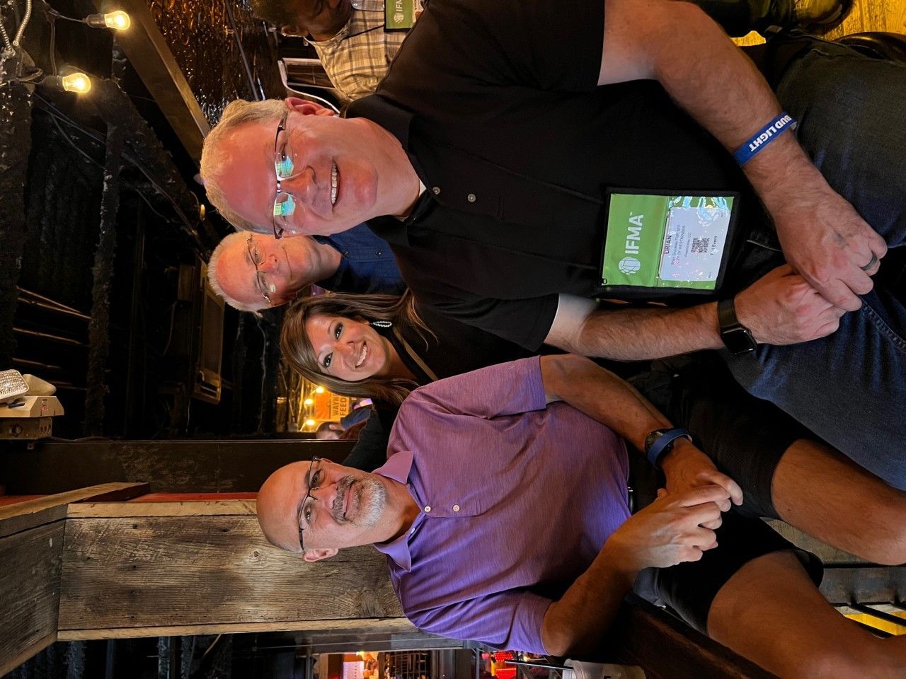 Group of people smiling, indoors. Two men in foreground, others behind them. Dark setting, some wearing badges.