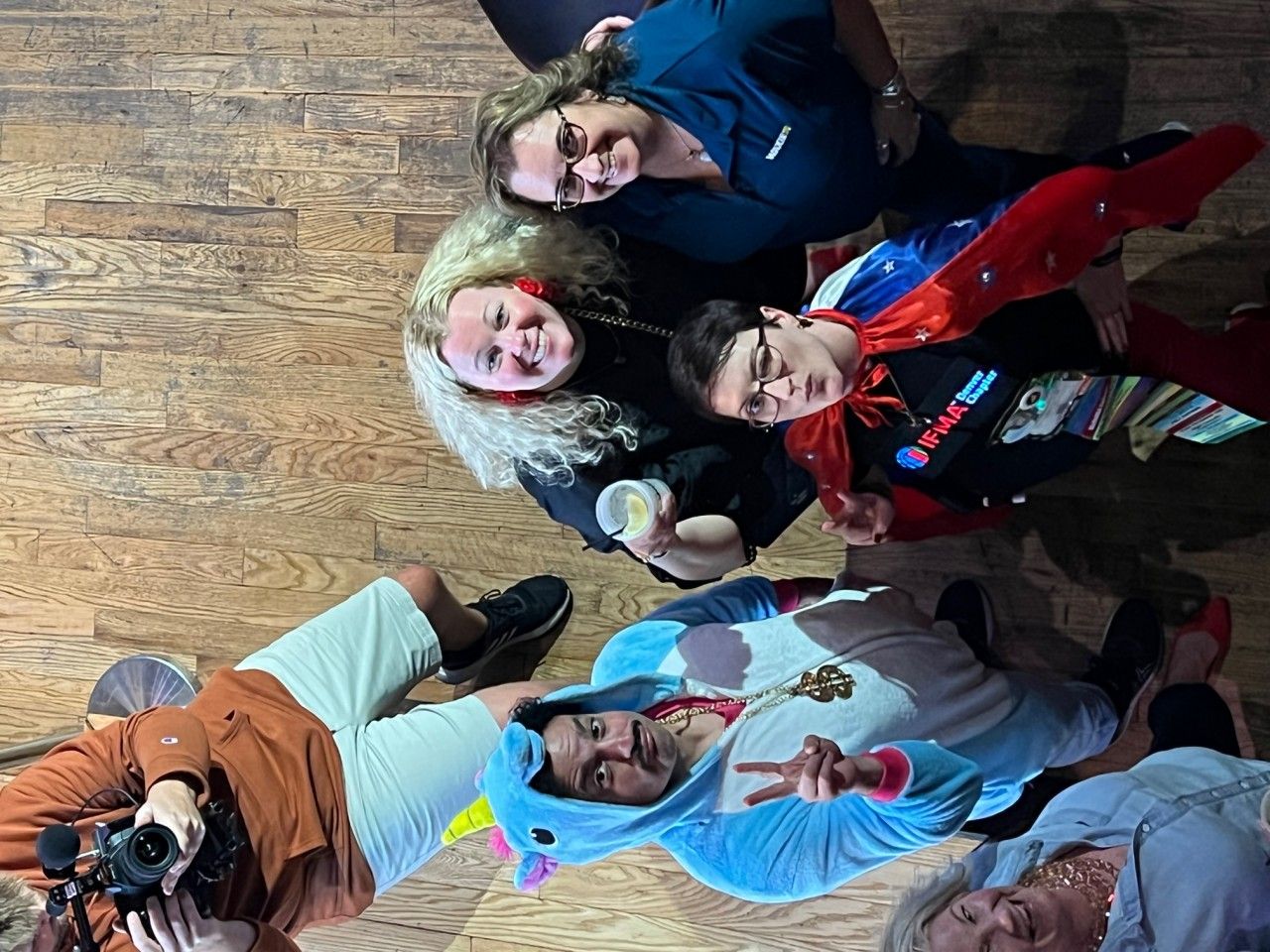 Group of people on a wooden floor, one in unicorn costume, one in cape, others smiling at camera.