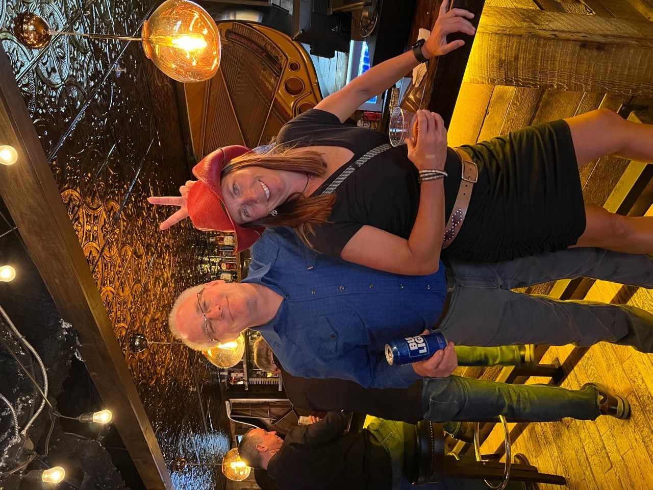 Two people smiling, posing in a bar. Woman in black dress, man in blue shirt, holding a can, and a man at a table.