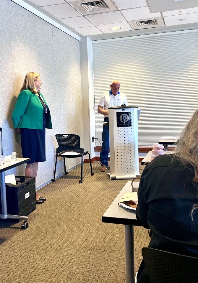 A man speaking at a podium in a conference room, a woman stands to the side.