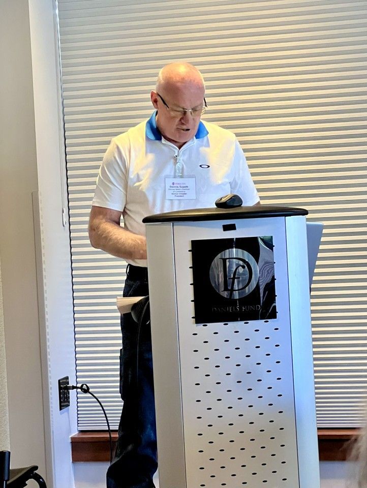 Man speaking at a podium with a logo, white shirt, and blue collar in a room with blinds.