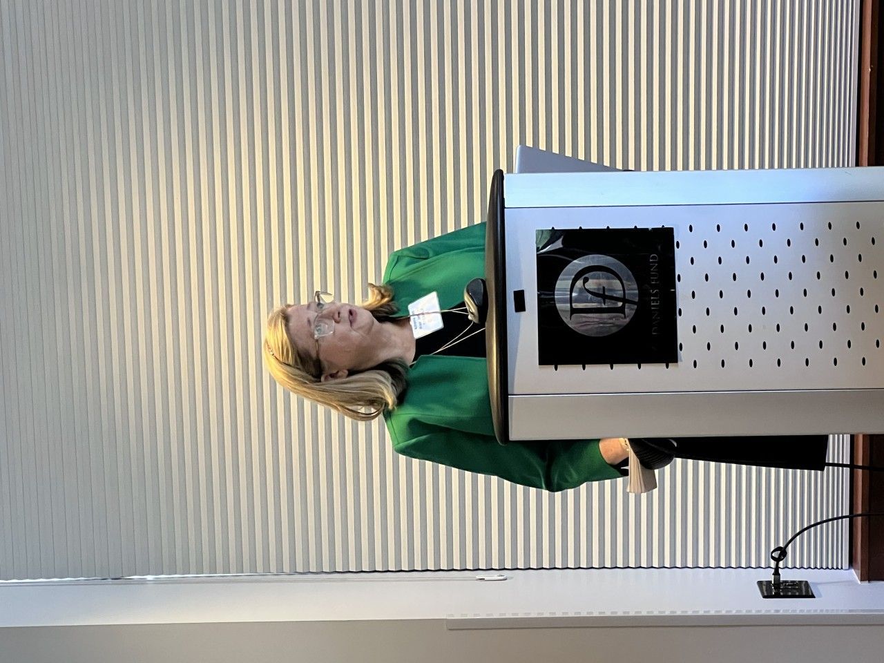 Woman in green jacket speaking at a podium, light-colored background.