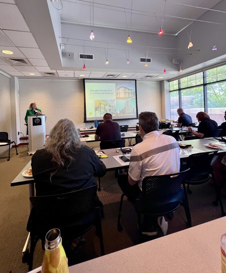 A presentation in a meeting room: speaker at podium, audience seated at tables, projected image on screen.