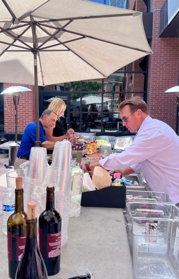 Outdoor bar setup with people. Man pours drink. Wine bottles, cups, and food items on the bar.