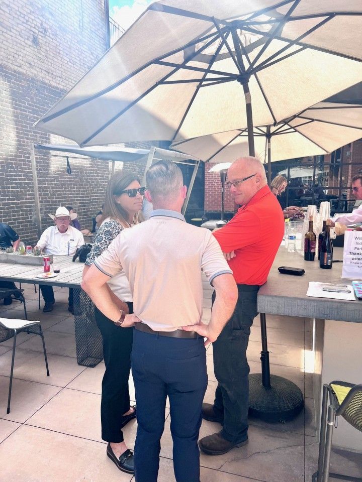 People conversing outdoors at a bar, under umbrellas. One man in an orange shirt. Sunny day.