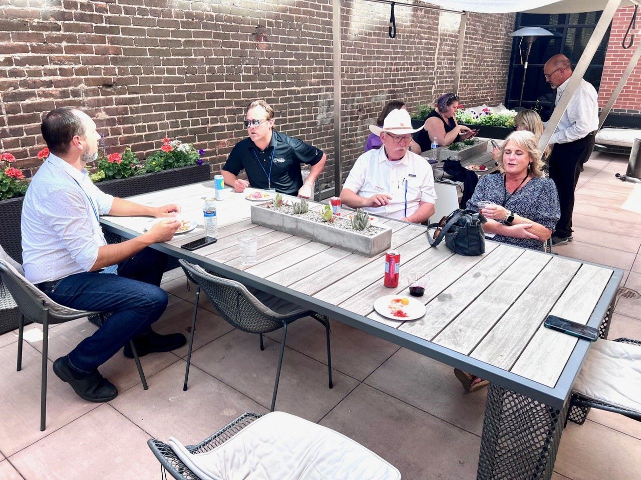People seated at an outdoor table with food and drinks; brick wall backdrop; sunny day.