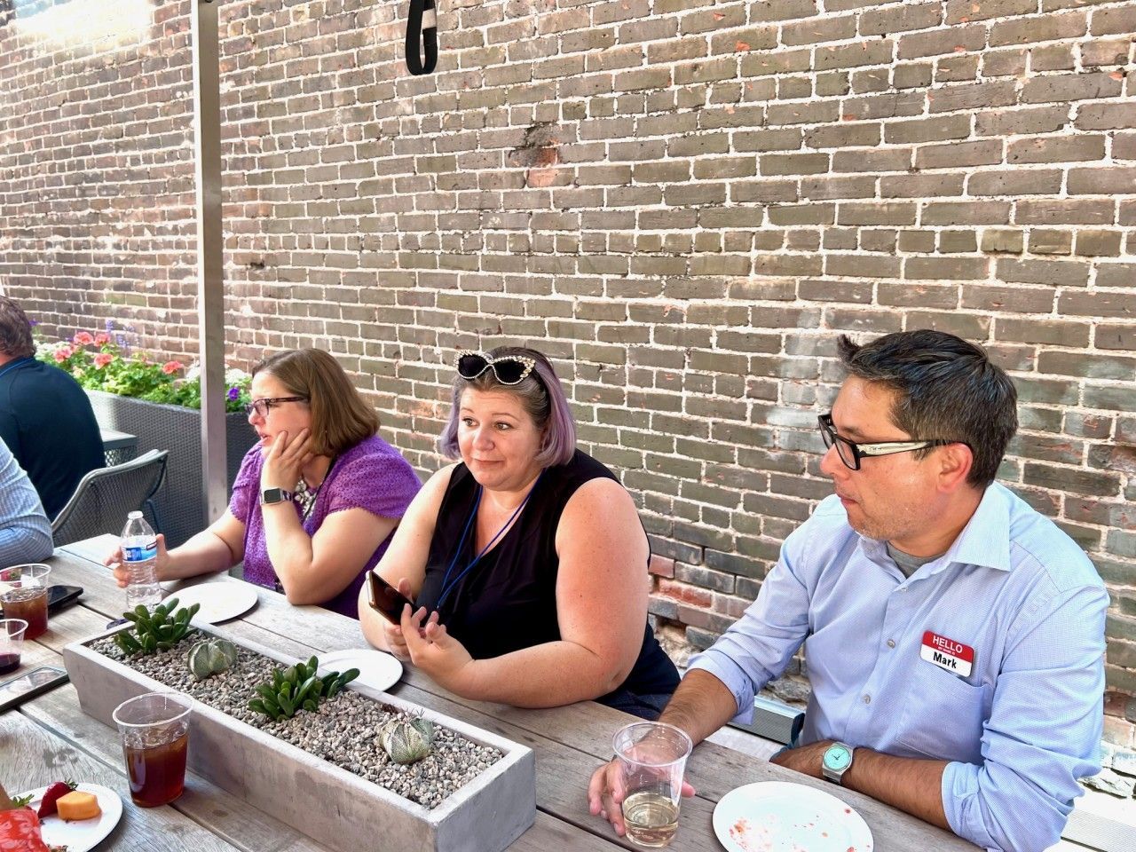People at an outdoor table; brick wall background. Man with glasses and name tag gestures, two women look on.