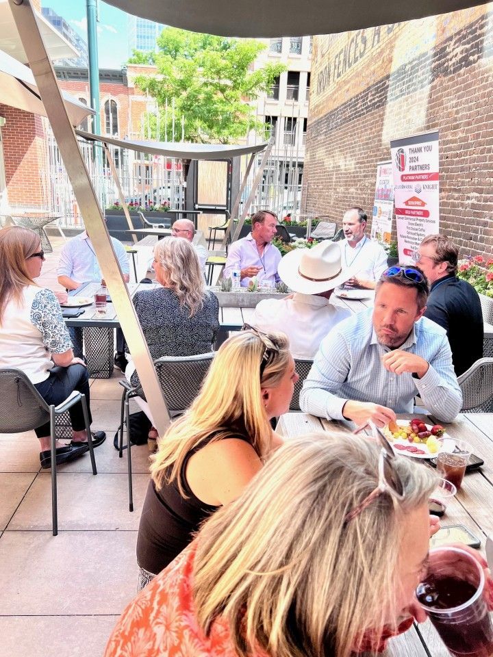 People at outdoor patio tables, enjoying drinks and food. Buildings, trees in background, sunny day.