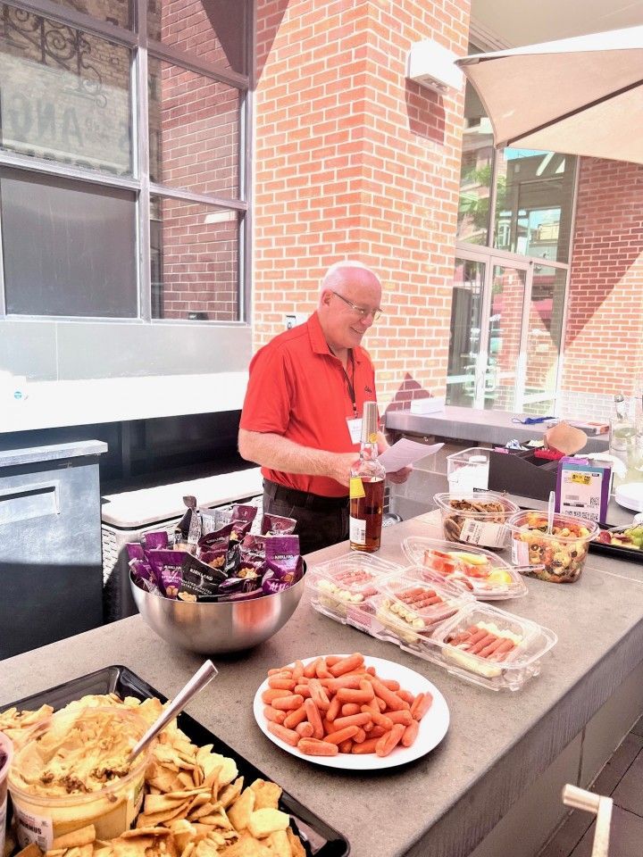 Man in red shirt prepares food at a buffet table, snacks include chips, hot dogs, and sandwiches; brick building background.