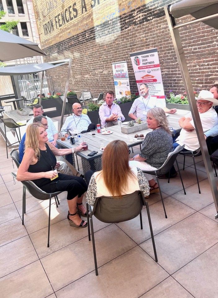 People seated around an outdoor table, some talking. A brick wall and banner are in the background.