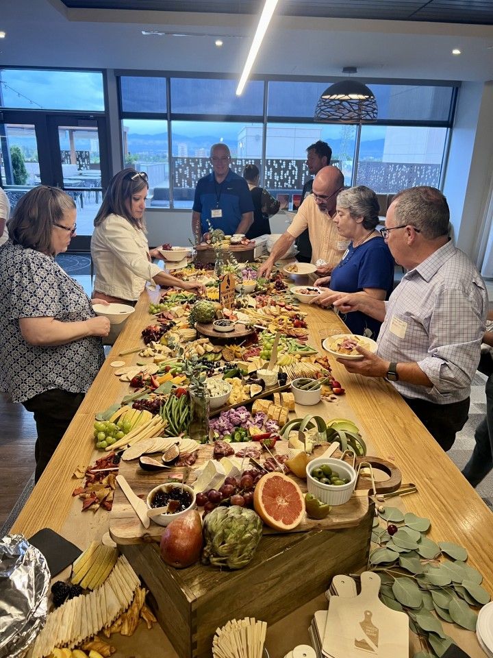 People at a long table with a charcuterie board. Windows overlook water.