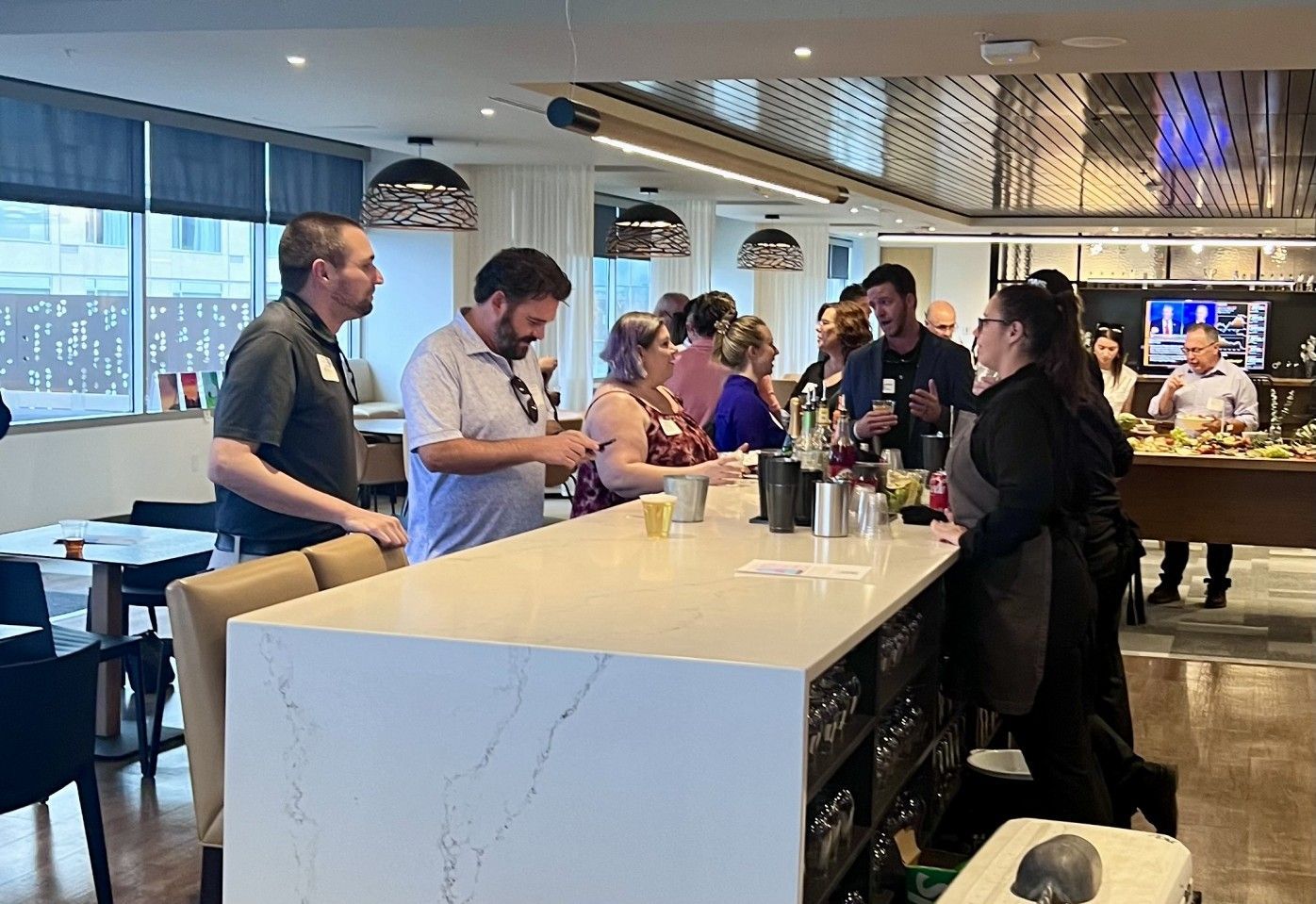 People at a bar, some talking, others near bartender; bright interior with snacks and drinks.