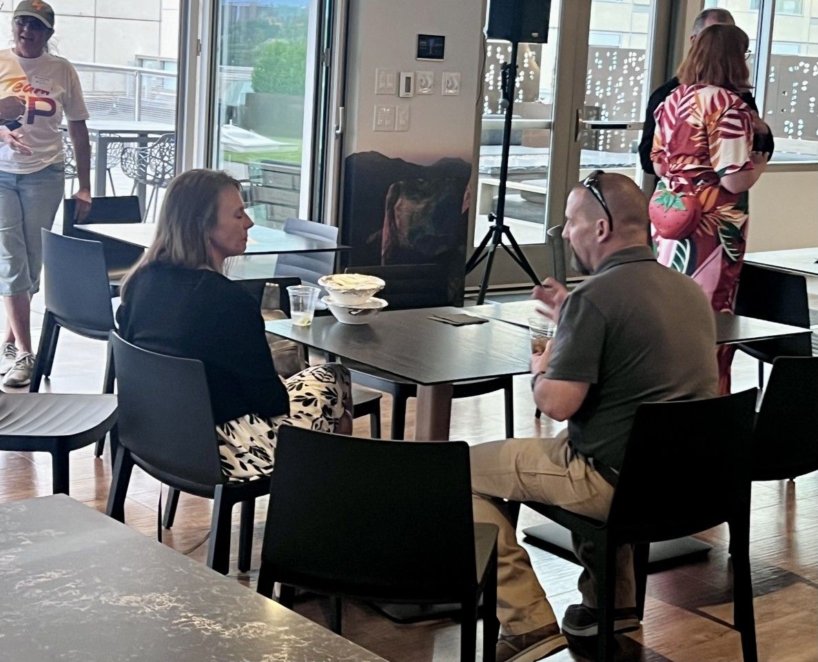 People seated at tables in a room, conversing. Some are standing, near a door and window.