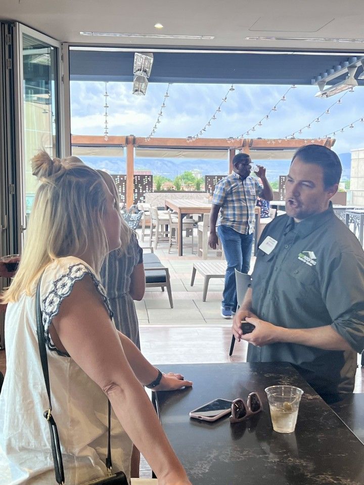 A man in a green shirt converses with a woman in front of a bar, outdoor seating in background.
