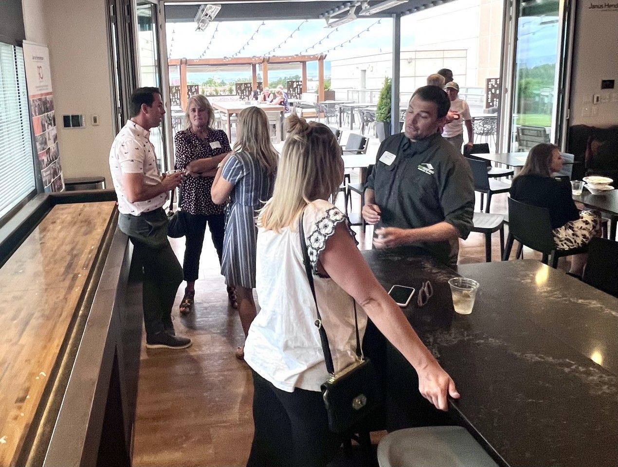 People chatting in a bar, some near a bar counter. A server is taking an order. Outside, a patio with more people.