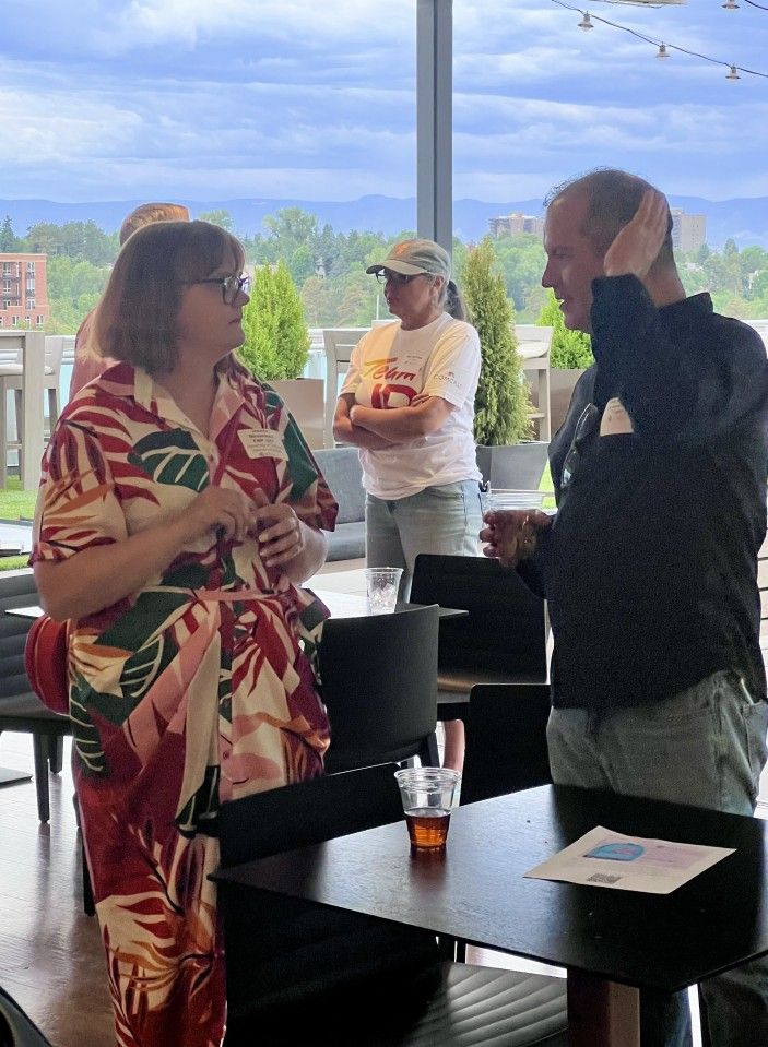 Three people conversing on a patio with city and mountain views. One wears a floral shirt, one holds a drink.