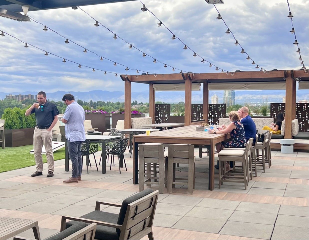 People on a rooftop patio with seating, tables, and string lights, with a mountain view.