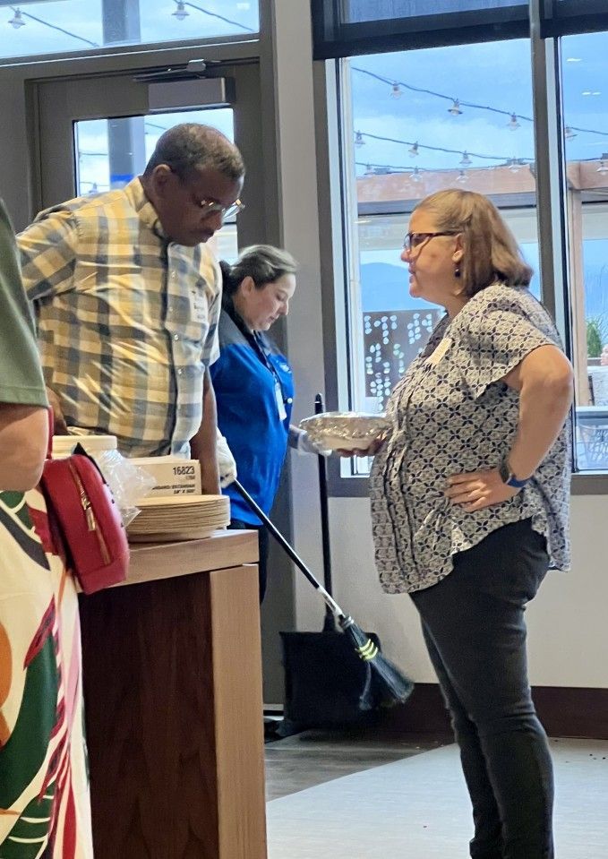 People conversing indoors near a table with food. Woman in patterned top holds a tray.