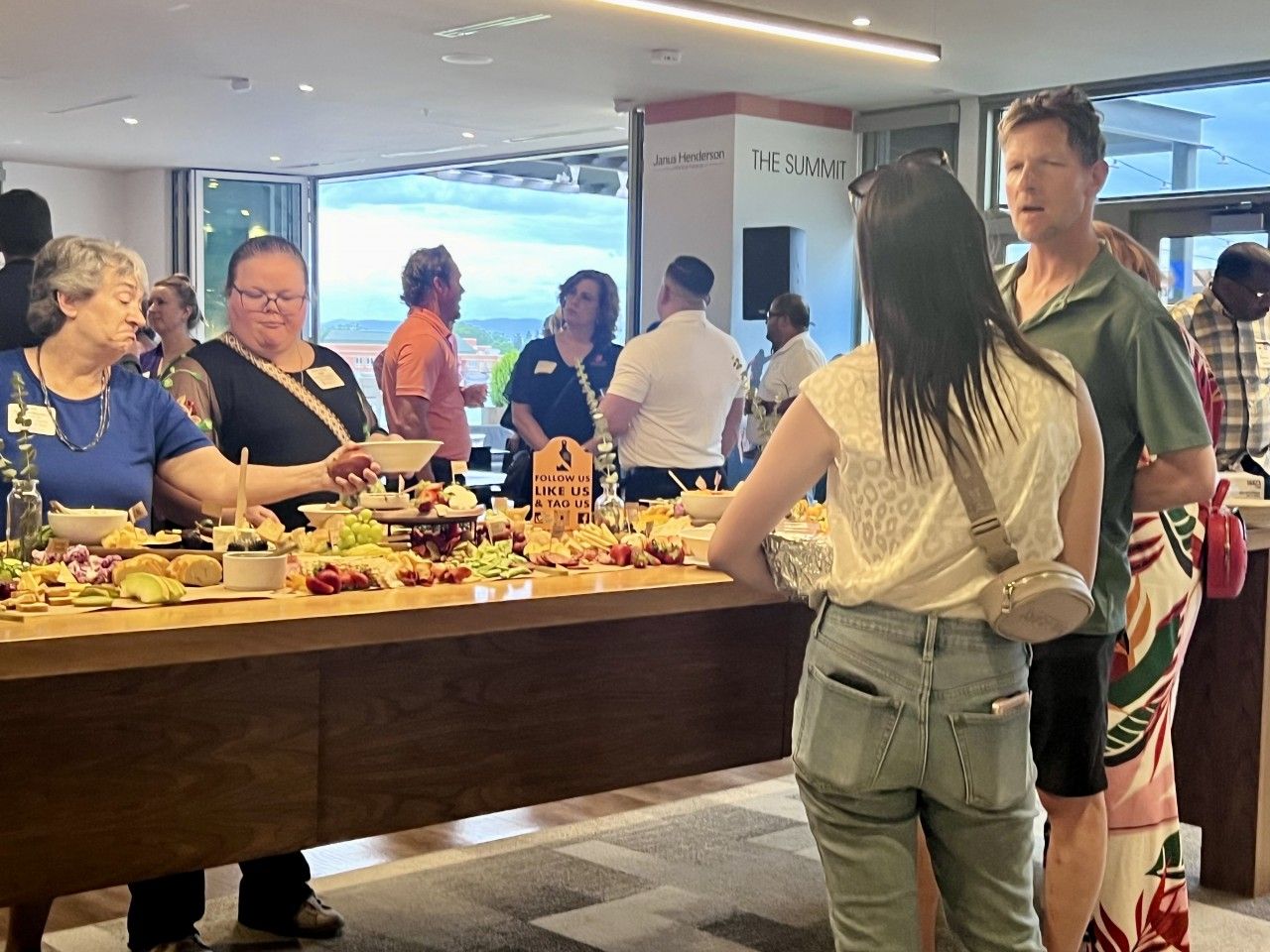 People at a buffet table, an event. Various food items on display. Indoors, open space, window with a view.