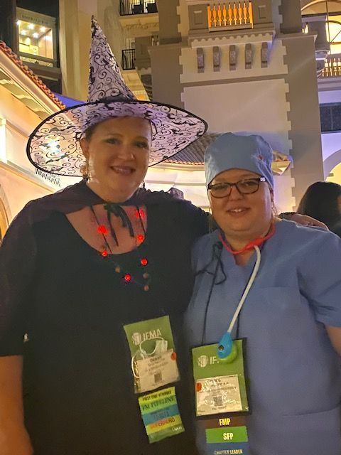 Two people in costumes, a witch and a medical professional, smiling, outdoors at night, wearing lanyards.