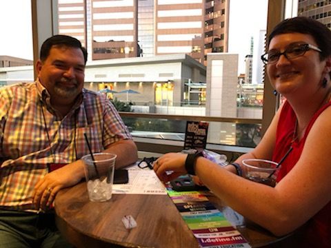 Two people seated at a table on a patio, smiling. City buildings are visible in the background.