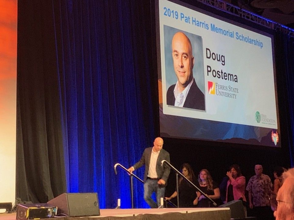 Man on stage accepts scholarship award. Screen shows Doug Postema's photo. People applaud.