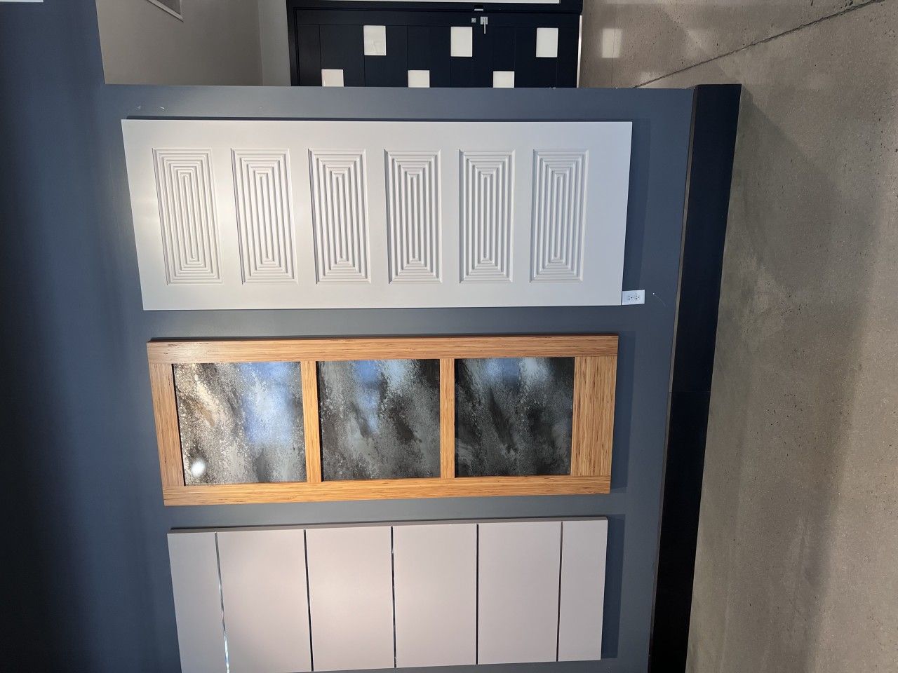 Three different door panel samples displayed on a blue wall: white with patterns, wood-framed with glass, and plain white.