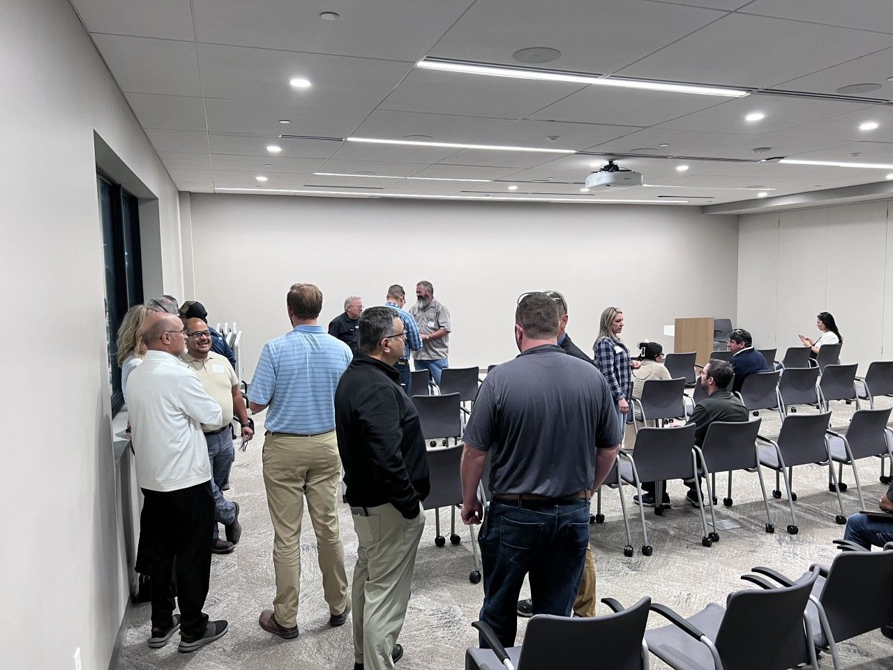 People in a brightly lit meeting room, some standing and talking, others seated in chairs.