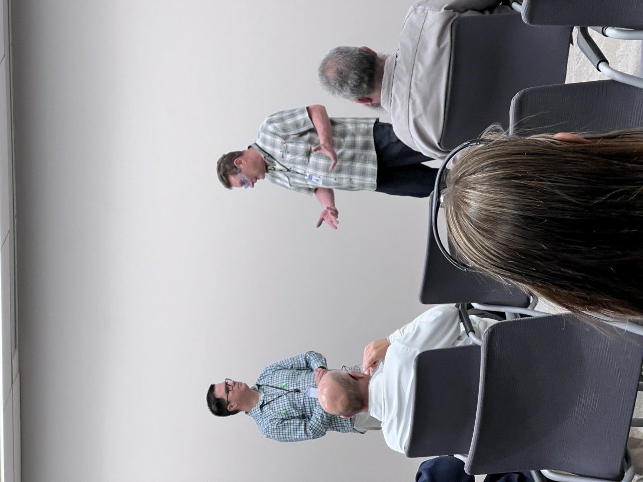 A man gestures while speaking to others seated in chairs. Indoors, neutral background.